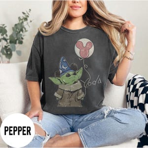 K&ouml;nnte beinhalten: Dunkelgraues T-Shirt mit einer gr&uuml;nen Cartoonfigur, die einen blauen Zaubererhut tr&auml;gt und einen rosa Mickey Mouse-Ballon h&auml;lt. Das Wort "Yoda" ist in einer Schreibschrift geschrieben. Das Wort "PEPPER" ist unten in einem wei&szlig;en Feld aufgedruckt.
