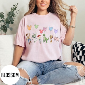 K&ouml;nnte beinhalten: Hellrosa T-Shirt mit einer Grafik von Zeichentrickfiguren, die Mickey Mouse-f&ouml;rmige Ballons in verschiedenen Farben halten. Das Wort "BLOSSOM" ist in einem wei&szlig;en Kreis unten links aufgedruckt.