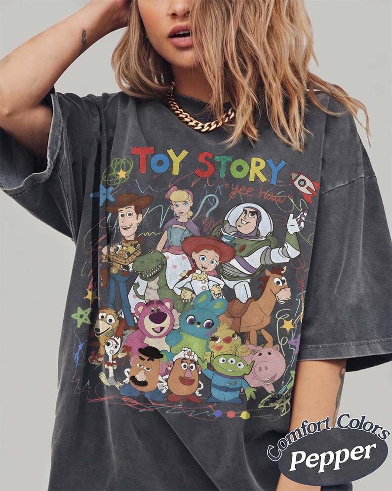May include: A charcoal gray short-sleeve t-shirt featuring a colorful graphic print of Toy Story characters including Woody, Buzz Lightyear, Jessie, and others. The design includes the text "TOY STORY" and "yee haw" with scribbled accents.