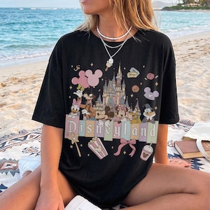 May include: A black short-sleeved t-shirt featuring a graphic print of a castle, Mickey Mouse, Minnie Mouse, Donald Duck, Daisy Duck, Goofy, and Pluto. The design includes the text Disneyland, pink balloons, popcorn, a bow, and stars.