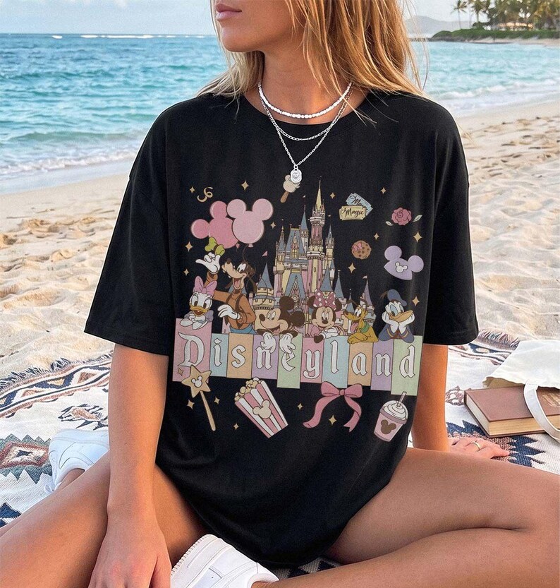 May include: A black short-sleeved t-shirt featuring a graphic print of a castle, Mickey Mouse, Minnie Mouse, Donald Duck, Daisy Duck, Goofy, and Pluto. The design includes the text Disneyland, pink balloons, popcorn, a bow, and stars.