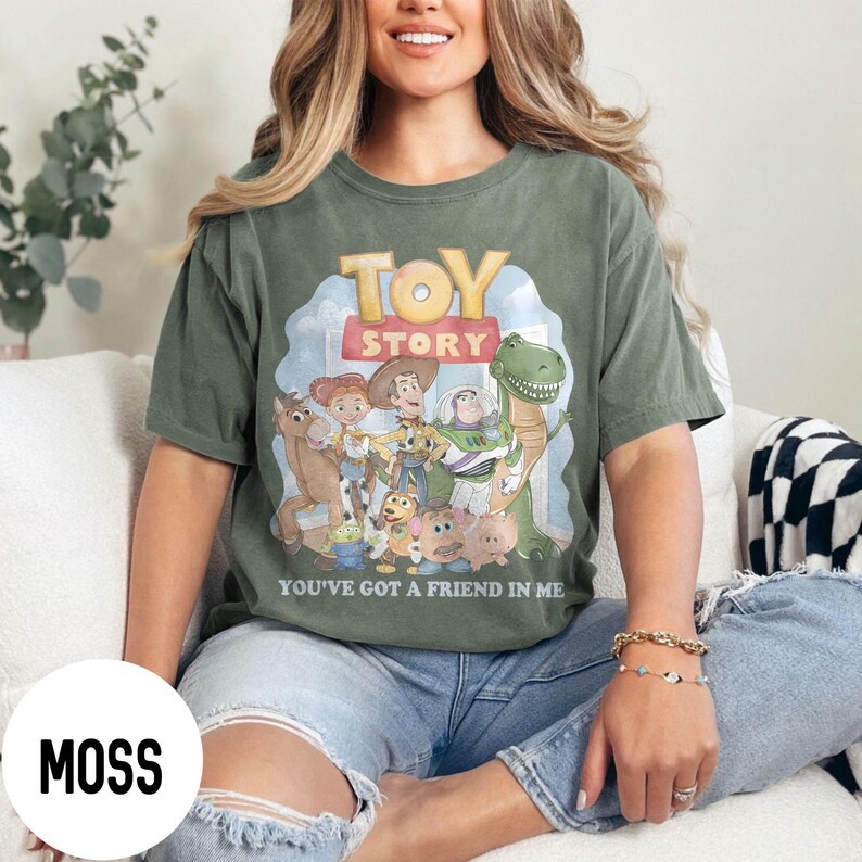 K&ouml;nnte beinhalten: Moosgr&uuml;nes T-Shirt mit den Worten "TOY STORY" in Gelb und Rot, sowie einer Grafik von Figuren aus dem Film. Der Satz "YOU'VE GOT A FRIEND IN ME" ist darunter gedruckt. Das T-Shirt wird von einer Person getragen.