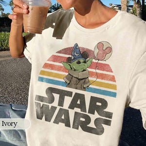 May include: Ivory-colored Star Wars t-shirt featuring a graphic of Grogu wearing a wizard hat and holding a Mickey Mouse balloon. The design includes a colorful sunset and the Star Wars logo in large, distressed letters.