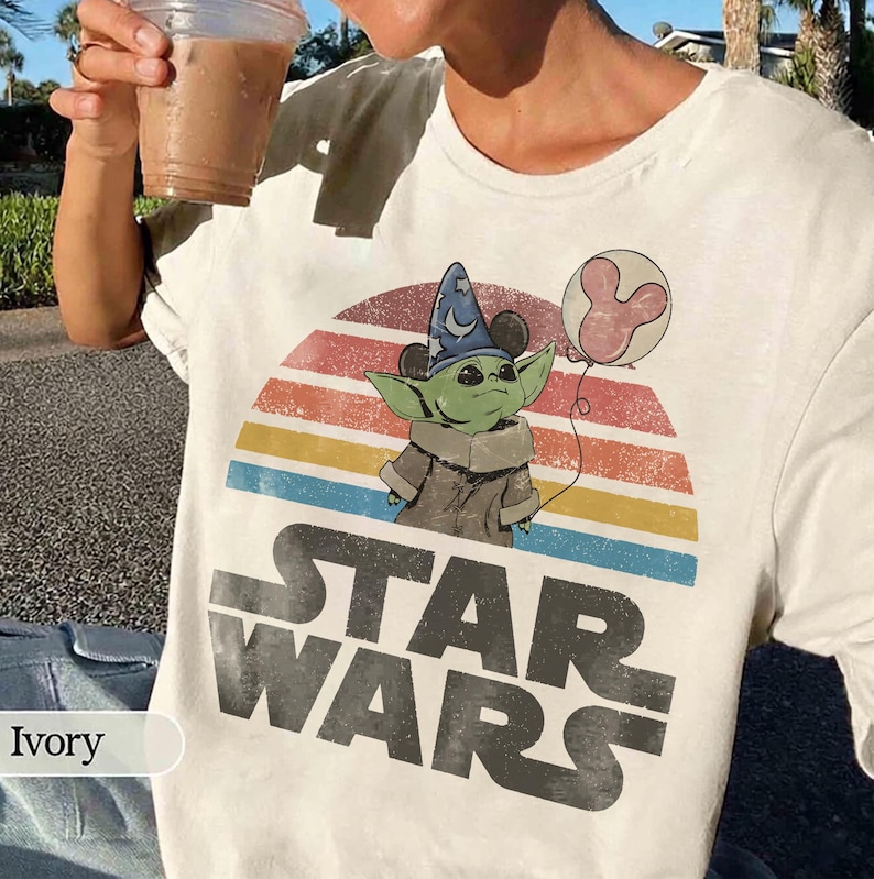 May include: Ivory-colored Star Wars t-shirt featuring a graphic of Grogu wearing a wizard hat and holding a Mickey Mouse balloon. The design includes a colorful sunset and the Star Wars logo in large, distressed letters.