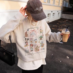 May include: A beige sweatshirt featuring Disney characters and landmarks, with the text "Walt Disney World" and "2026". The sweatshirt is paired with a brown baseball cap that says "La Paris". A black purse and a beverage are also visible.