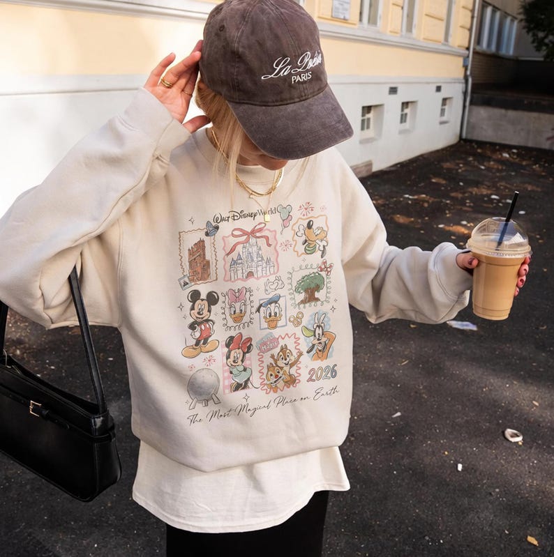 May include: A beige sweatshirt featuring Disney characters and landmarks, with the text "Walt Disney World" and "2026". The sweatshirt is paired with a brown baseball cap that says "La Paris". A black purse and a beverage are also visible.