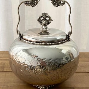 May include: A silver-colored cracker jar with a lid and handle. The jar has floral engravings and the word "CRACKERS" etched on the side. The handle and lid finial feature ornate detailing. The jar sits on a wooden surface.