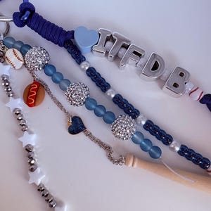 May include: A keyring featuring a blue carabiner, dark blue paracord, and silver letter beads spelling ITFDB. The design includes blue matte beads, silver rhinestone spheres, white star beads, and charms shaped like a baseball, hot dog, and heart.