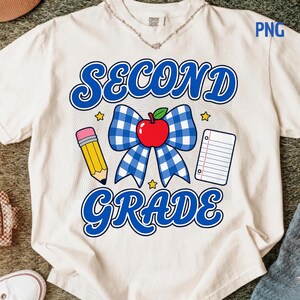 May include: A cream-colored t-shirt featuring a graphic with the text SECOND GRADE in blue. The design includes a blue and white gingham bow, a red apple, a yellow pencil, a piece of lined notebook paper, and small yellow stars on a background.