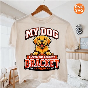 May include: Cream-colored t-shirt featuring a cartoon golden retriever with the text "MY DOG" above and "PICKED THE PERFECT BRACKET" below. The dog is smiling, and the design includes a basketball.