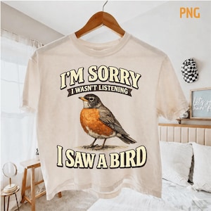 May include: Cream-colored t-shirt with the text "I'M SORRY I WASN'T LISTENING I SAW A BIRD" and a detailed illustration of a robin. The bird has orange, brown, and gray feathers. The shirt is hanging on a wooden hanger.