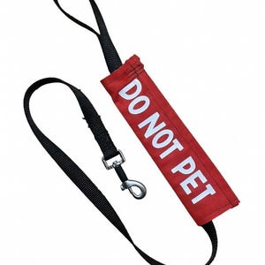 May include: A black dog leash with a red fabric handle. The handle has the white text "DO NOT PET". The leash has a metal clasp and a loop handle.