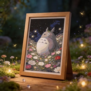 Puede incluir: Un bordado de punto de cruz enmarcado con un personaje Totoro gris en un campo de flores coloridas bajo un cielo estrellado. El marco de madera está apoyado sobre una superficie de madera, con un fondo borroso de flores y luces.