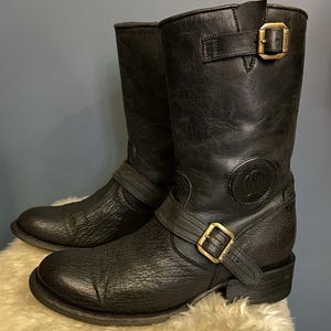 Könnte beinhalten: Ein Paar wadenhohe Stiefel aus schwarzem Leder mit strukturierter Oberfläche am Fußbereich und glatterem Lederschaft. Jeder Stiefel verfügt über einen messingfarbenen Schnallenriemen am Knöchel und eine passende Schnalle oben sowie ein Logo.