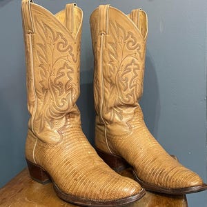 Könnte beinhalten: Ein Paar hellbraune Leder-Cowboystiefel mit einer strukturierten Eidechsenhaut-Optik am Fußteil und einem glatten Lederschaft mit aufwendigen Ziernähten. Die Stiefel haben eine spitz zulaufende Kappe, einen Absatz aus Holz und Anziehschlaufen.