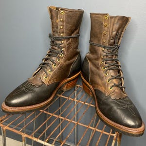 Pode incluir: Um par de botas de cowboy de couro castanho e preto. As botas têm um design com atacadores com ilhós de latão e um padrão costurado decorativo na biqueira. As botas estão numa prateleira de metal enferrujado.