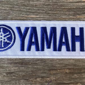 May include: A rectangular white patch with the word "YAMAHA" embroidered in dark blue capital letters. The patch also features a blue and white emblem on the left side. The patch is set against a wooden background.