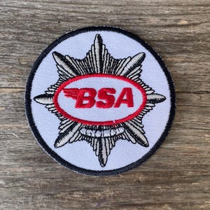 Puede incluir: Parche bordado con fondo blanco, con un óvalo rojo con las letras "BSA" y un diseño de ala estilizado. El óvalo está rodeado por un diseño de estrella en blanco y negro, con un borde negro.