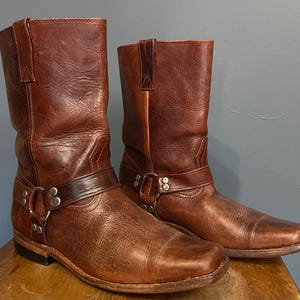 Puede incluir: Un par de botas de cuero marrón a media pantorrilla con punta cuadrada, tacón de bloque y una correa de arnés decorativa en el tobillo. El arnés está asegurado con anillos y remaches de metal plateado, y el cuero tiene un acabado texturizado.