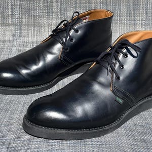 以下が含まれることがあります： 黒い革のチャッカブーツ一足。黒い靴紐が付いています。ブーツは丸いつま先と厚い黒いソールです。内側は明るい茶色です。ブーツは灰色のテクスチャのある表面に表示されています。