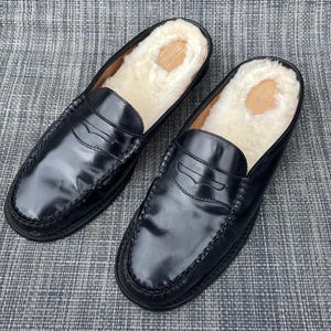 May include: Pair of black leather slip-on loafers with a glossy finish. The interior is lined with a soft, cream-colored shearling material. The shoes have a classic penny loafer design.