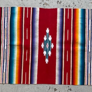 Könnte beinhalten: Ein rechteckiges gewebtes Textil mit einem lebendigen Streifenmuster in Rot, Grün, Gelb, Orange, Blau und Violett. Die Mitte zeigt ein rautenförmiges geometrisches Motiv in Weiß und Türkis. Die Enden sind mit langen weißen Fransen versehen.