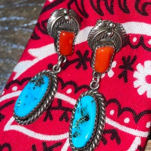 Może przedstawiać: Srebrne kolczyki z turkusowymi i koralowymi detalami. Każdy kolczyk ma górę w kształcie liścia, jasnopomarańczowy kawałek koralowca i owalny turkusowy kamień ze srebrną obwódką ze sznurka. Kolczyki spoczywają na czerwono-białej tkaninie we wzory.