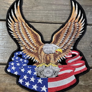 May include: Embroidered patch featuring a majestic eagle with outstretched wings, a motorcycle engine, and a banner with the American flag design. The eagle has brown and white feathers, a yellow beak, and a gray head. The banner has red, white, and blue colors.