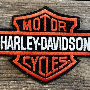 May include: Embroidered Harley-Davidson patch in black, orange, and white. The patch features the words "MOTOR HARLEY-DAVIDSON CYCLES" in a shield-shaped design. The patch is likely for a jacket or vest.