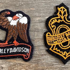 May include: Two Harley-Davidson embroidered patches. One shows a brown and white eagle with the text "HARLEY DAVIDSON" on a banner. The other patch is black with gold embroidery and the text "HARLEY DAVIDSON MOTOR CO."