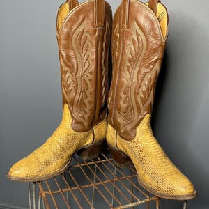 Op de afbeelding: Een paar cowboylaarzen met een bruin leren bovenwerk en een gele slangenleerachtige voorkant. De laarzen hebben decoratieve stiksels en staan op een metalen rek. Een klassieke Western-stijl.