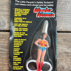 Könnte beinhalten: Vintage Snippys Sicherheitsschere mit Wonder Woman-Design. Die Schere hat weiße Griffe und eine silberne Klinge, mit einer Wonder Woman-Figur oben. Die Verpackung ist schwarz mit rotem, gelbem und blauem Text.