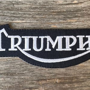 Könnte beinhalten: Schwarzer, gestickter Aufnäher mit dem weißen Wort "TRIUMPH" und einem stilisierten weißen Bogen darunter. Der Aufnäher hat eine einzigartige Form und befindet sich auf einem Holzgrund.