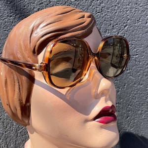 May include: Large, brown tortoiseshell sunglasses with a square frame. The sunglasses have a light brown tint and are set on a mannequin head with brown hair and red lipstick.