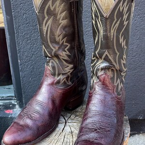 Könnte beinhalten: Ein Paar dunkelbraune Leder-Cowboystiefel mit einem strukturierten, tiefburgunderroten Vorderblatt und einem hohen Schaft mit aufwendigen, hellen Ziernähten. Die Stiefel haben eine spitz zulaufende Kappe, einen Blockabsatz und Zuglaschen.
