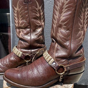 Può includere: Un paio di stivali da cowboy in pelle marrone scuro con gambali alti e complessi motivi di cuciture chiare. Ogni stivale è decorato con un cinturino rimovibile che presenta accenti color argento metallizzato e un piccolo ciondolo pendente.