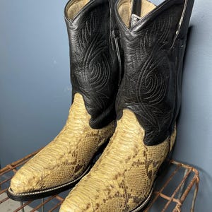 May include: A pair of cowboy boots with black leather uppers and tan snake-skin patterned vamps. The boots have a classic western design with decorative stitching and a pointed toe. The boots are displayed on a metal rack.