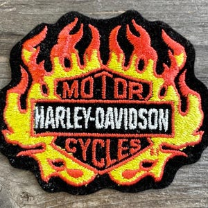May include: Embroidered Harley-Davidson patch with a black border and a flame design in yellow and red. The patch features the words "MOTOR HARLEY-DAVIDSON CYCLES" in white lettering.
