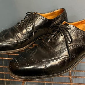 May include: A pair of black leather Oxford shoes with decorative perforations and lace-up closures. The shoes are displayed on a metal rack, showcasing their classic design and polished finish.