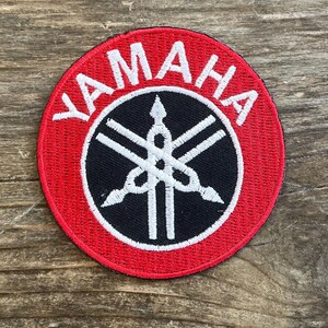 May include: Round, embroidered patch with the word "YAMAHA" in white capital letters on a red background. The center features a black circle with a white logo. The patch is likely for a motorcycle or musical instrument.