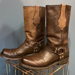 May include: A pair of brown leather motorcycle boots with a square toe and harness detail. The boots have a two-tone design, with a lighter brown upper and a darker brown lower section. The boots are displayed on a metal rack.