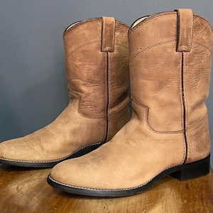 Op de afbeelding: Een paar halflange laarzen van beige leer met een ronde neus, treklussen aan de bovenkant van de schacht en donkerbruine biezen langs de naden. De laarzen hebben een zwarte zool en een lage blokhak met een matte, verweerde uitstraling.