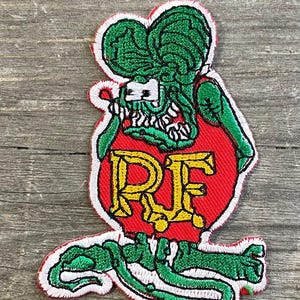 Op de afbeelding: Geborduurde patch met een cartoonrat met een groene kop en ledematen, een rode body met gele letters "RF" en een witte omtrek. De rat heeft een open mond en houdt een moersleutel vast. De patch ligt op een houten oppervlak.