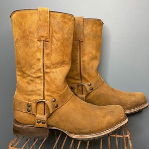 Puede incluir: Un par de botas de vaquero de cuero marrón. Las botas tienen un acabado envejecido, una altura media de la pantorrilla y una correa de arnés con herrajes metálicos. Las suelas son de color marrón claro con un aspecto desgastado. Las botas se exhiben en un estante de metal.