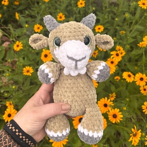 May include: A crochet plush toy shaped like a goat, featuring a tan body, a white muzzle, and gray accents on the horns and hooves. The toy has large, dark, round eyes and a soft, textured yarn finish, crafted with a detailed, stuffed construction.