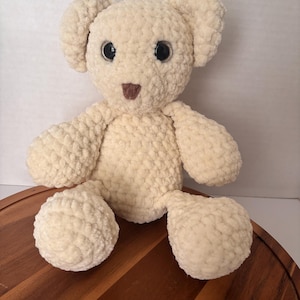 May include: A cream-colored, handmade crochet teddy bear with large black eyes and a small brown nose. The bear has rounded ears, arms, and legs, and is sitting upright. The plush toy is made of soft, textured yarn.
