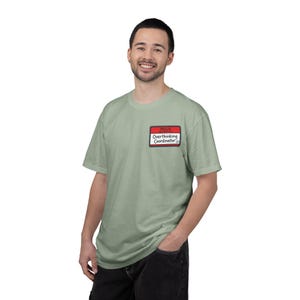 May include: A pale green crew neck t-shirt featuring a red and white name tag that says "HELLO. Overthinking Coordinator". The t-shirt is made of a soft material.