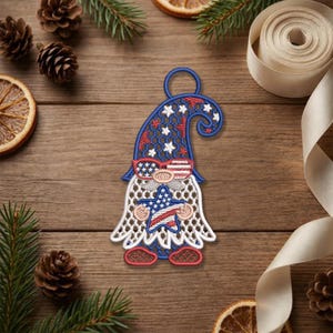 May include: Embroidered gnome ornament with a patriotic theme. The gnome wears a blue hat with white stars, sunglasses, and a star-shaped body with the American flag design. The ornament has a loop for hanging.