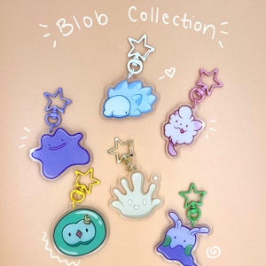 May include: A collection of colorful acrylic keychains featuring cartoon blob designs. Each keychain has a star-shaped clasp. The keychains are in various colors, including blue, purple, green, and white, with the words "Blob Collection" written above.