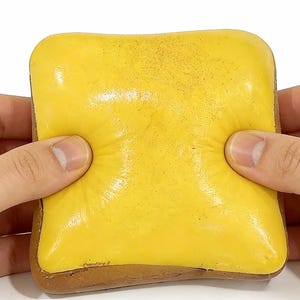 May include: A yellow, square-shaped stress toy resembling a slice of cheese, with a brown crust. The toy is being squeezed by two hands, showing the soft, pliable texture. The cheese slice is a vibrant yellow color.